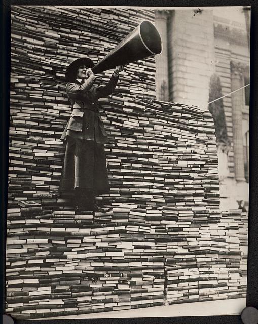 An old sepia toned photograph of a woman on a stack of books. She holds a megaphone.