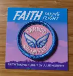 Photo of a blue and pink patch on a cardboard background. Patch has blue and white wings on a pink background and white lettering that says Landed on My Feet. It has a blue embroidered edge. The background has title information - Faith: Taking Flight above the patch, Faith: Taking Flight by Julie Murphy below it. The background in the middle is a pink purple and blue sunset seen, with a city Horizon below.
