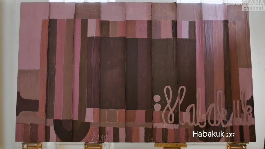A still from the Louisiana Channel film with Danish artist Tal R, showing one panel of nine from the series Habakuk. All nine are abstracted railcars, grand in scale, nearly life-sized. This one is done in tones of pinks and browns, and rests upon chairs and a small table. There is a colon on the right side of the car, with the word Habakuk written in cursive just past it.