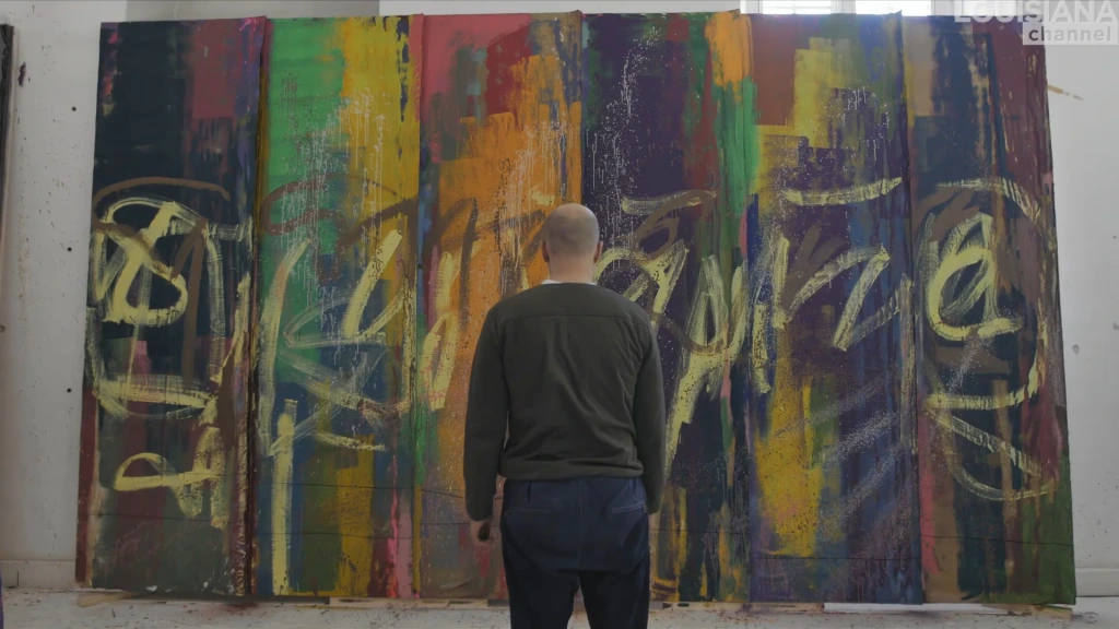 A still from the Louisiana Channel film with Danish artist Tal R, showing one panel of nine from the series Habakuk. All nine are abstracted railcars, grand in scale, nearly life-sized. This one is apparently in process, the artist seen from the back. In front of him, a multi-colored 'railcar' with yellowish markings reminiscent of graffiti arced across it.